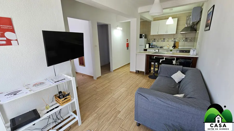 Sala com sofá cinzento, televisão e cozinha aberta ao fundo